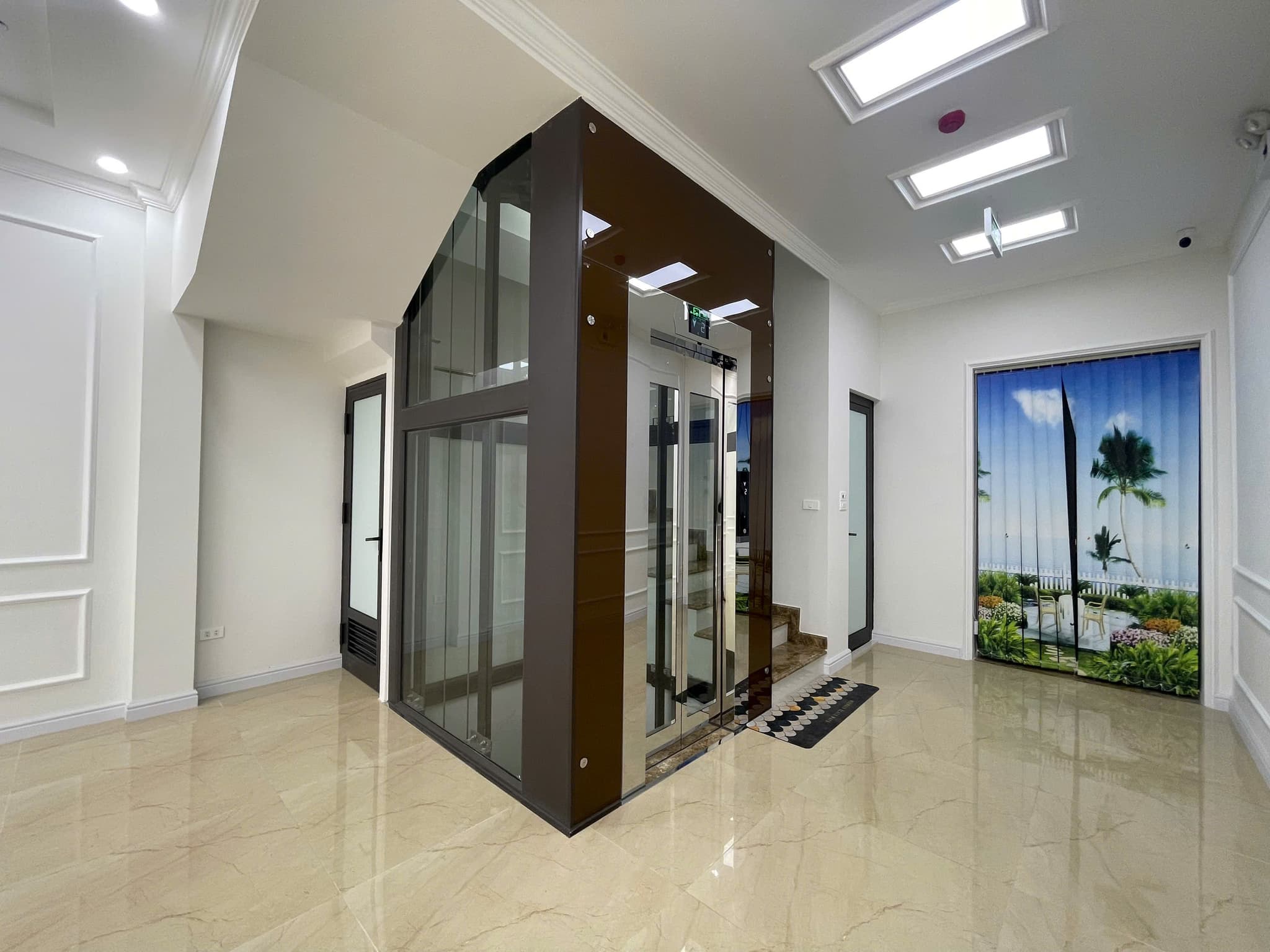 Thang máy kính cường lực 450kg sơn màu nâu trà, đặt giữa phòng khách lát đá marble sáng