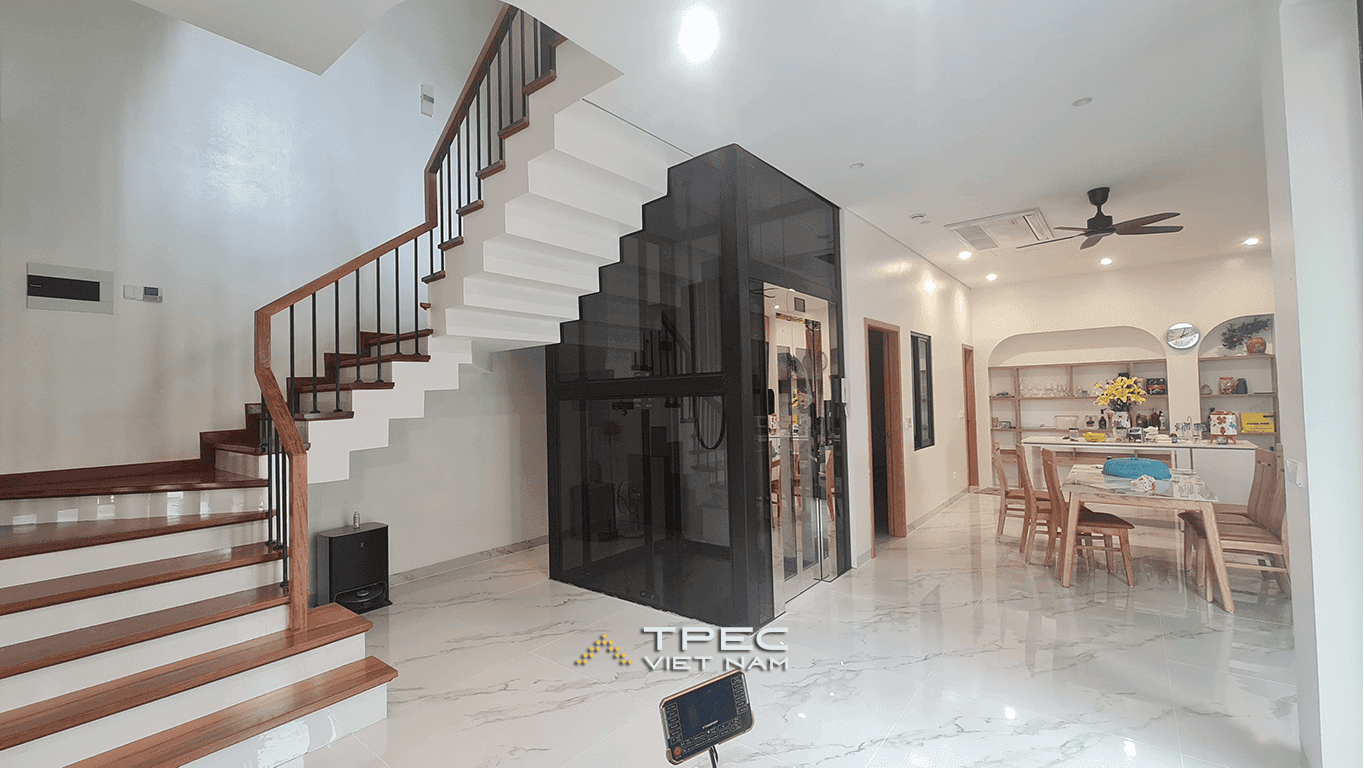 Không gian tầng 1 nhà phố Đông Anh với thang máy kính khung đen đặt cạnh cầu thang, liền phòng bếp