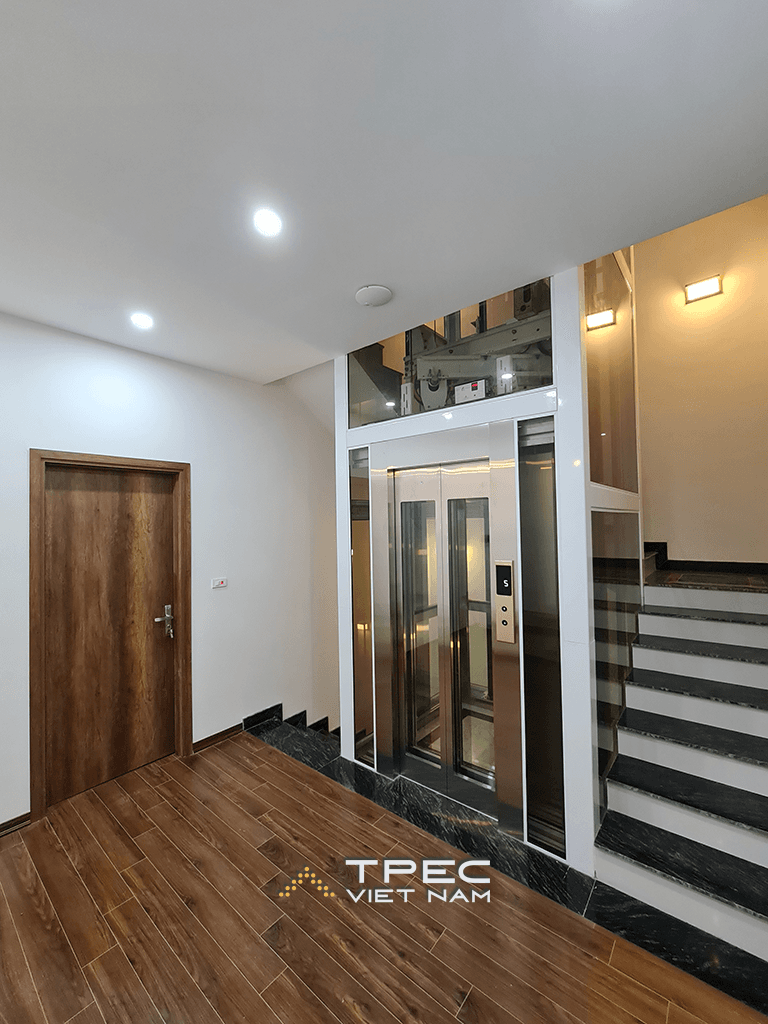 Toàn cảnh thang máy kính khung trắng đặt cạnh cầu thang trong nhà phố Him Lam Vạn Phúc