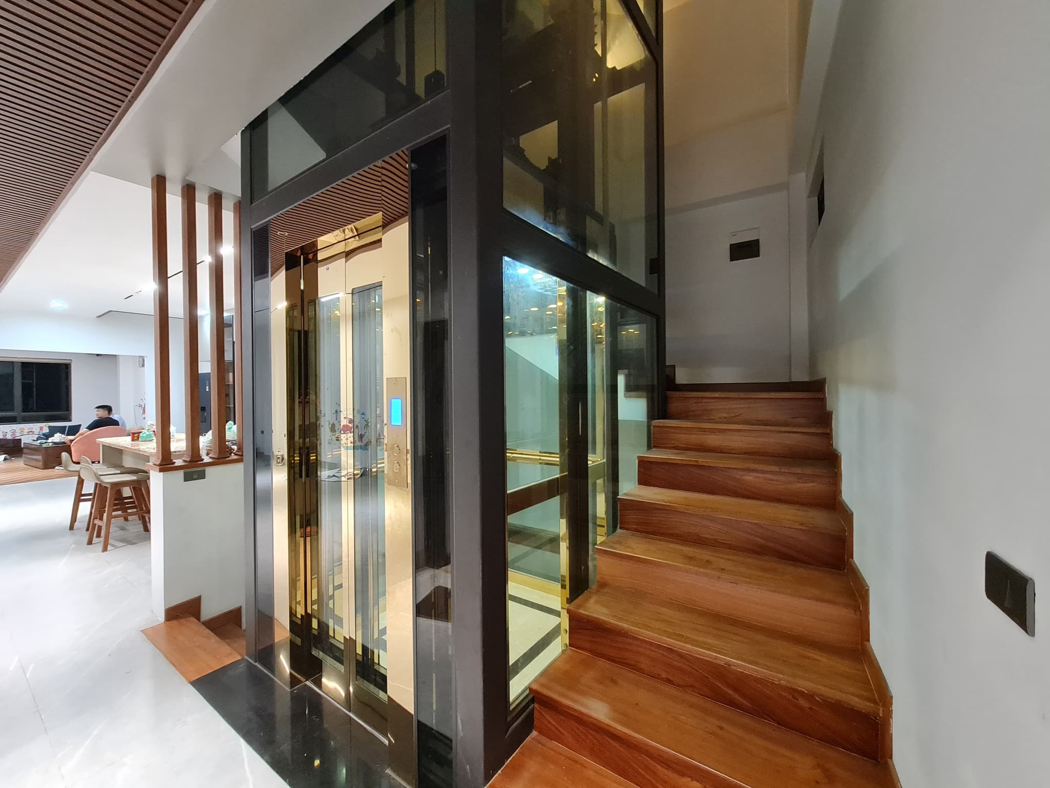 Thang máy kính 350kg giếng kính khung đen cạnh cầu thang gỗ, nhìn xuyên vách thấy cabin ánh vàng